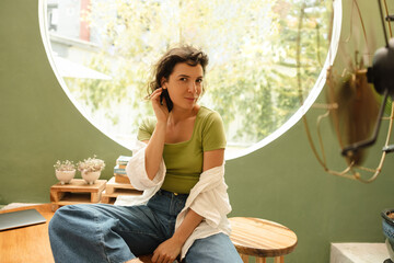 Beautiful young caucasian woman posing looking at camera, straightening her hair sit indoors. Brunette wears t-shirt, shirt and jeans. Lifestyle concept