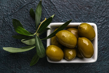 Bowl with tasty green olives and leaves on dark background