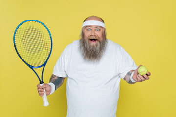 astonished plus size man with tennis ball and racquet looking at camera isolated on yellow.