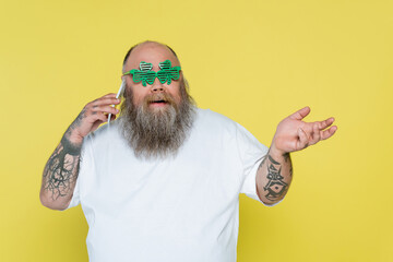 bearded overweight man in clover-shaped glasses talking on smartphone isolated on yellow.