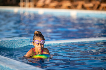 A girl in a bright bathing suit swims with an inflatable ball in a pool with clear water on a summer evening