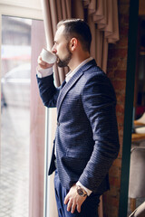 A young man in a business suit is drinking coffee by the window. A young man with a beard in a cafe.