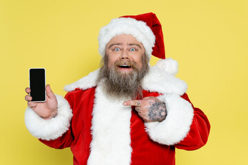 excited overweight santa claus pointing at cellphone with blank screen isolated on yellow.