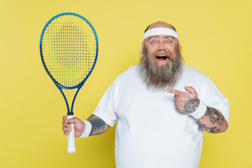 amazed plus size man pointing at tennis racquet and looking at camera isolated on yellow.