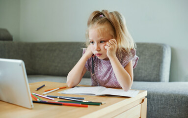 Distant education. Little girl is using tablet sitting at table. Female child having online classes via tablet computer at home