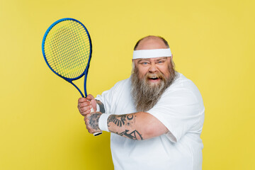thrilled overweight and tattooed man playing tennis isolated on yellow.