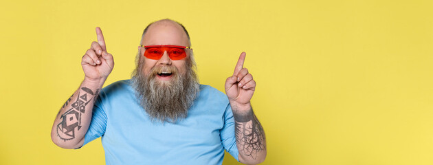 joyful bearded man with overweight looking at camera and pointing up with fingers isolated on...