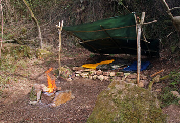 Bushcraft shelter built in the middle of an atlantic tree forest. Wooden shelter with a tarp and a fire pit made of stones. Campsite built outdoors and concept of survival activities.