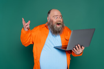 amazed overweight man with beard looking at laptop isolated on green.