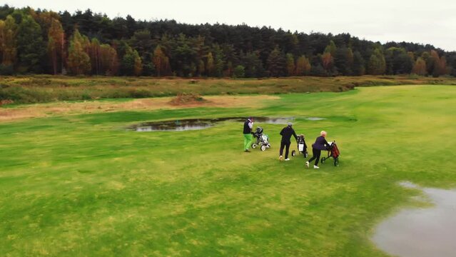 Sport concept. Three people with heavy big bags with golf eqiupment walking on a wet flat golf course, prepering for the next game. High quality 4k footage