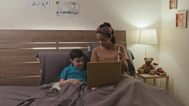 Asian boy and his mother discussing photos in family album while resting together in bed in evening at home