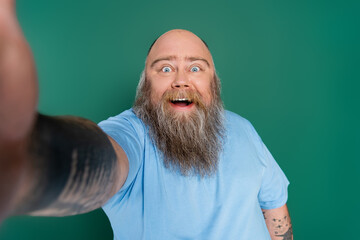 cheerful man with beard and overweight taking selfie on blurred foreground isolated on green.