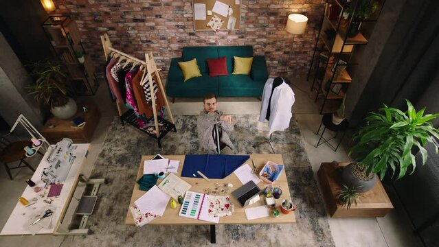Top view. A handsome man throws a perfect paper airplane into the air, he is working as a designer in his modern home office, measuring and cutting out a piece of blue fabric and he looks very