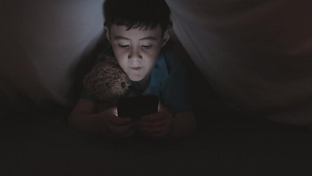 Little Asian boy lying on bed under blanket, hugging stuffed toy and browsing the Internet on smartphone