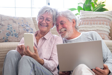 Cheerful caucasian senior couple sitting on the floor at home using laptop and smart phone, modern retired elderly people surfing the net with computer