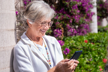 Portrait of beautiful elderly woman leaning on a plant while using the smart phone in a sunny summer day