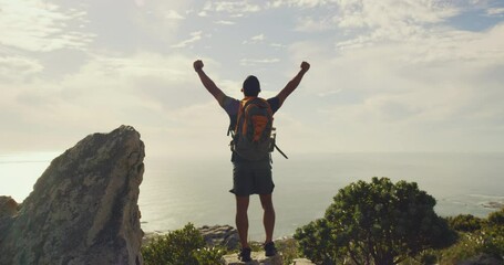 Young mountain hiker celebrating climbing to peak and cheering with fist and arms raised. Fit, athletic man admiring scenic view of ocean and sky. Backpacking alone in remote trekking area in nature
