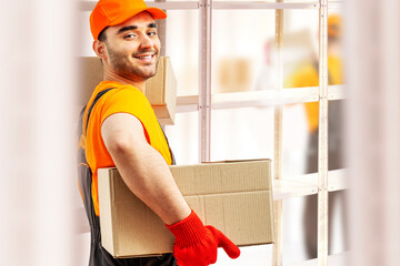 Young man holding cardboard package working in warehouse among racks and shelves. Delivery man with...