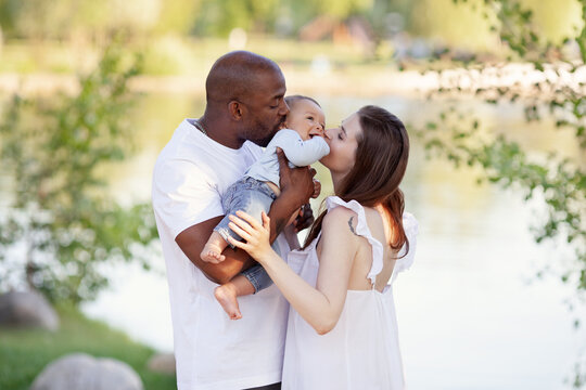 Happy Multiethnic family. Caucasian Mother and african american Father kiss small child. Parents, Portrait of Mom, dad and baby on hands over nature background