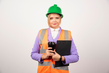Young female industrial engineer in uniform with clipboard and black cup on white background