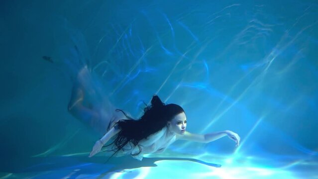 A girl in a white dress with long dark hair swims underwater as if floating