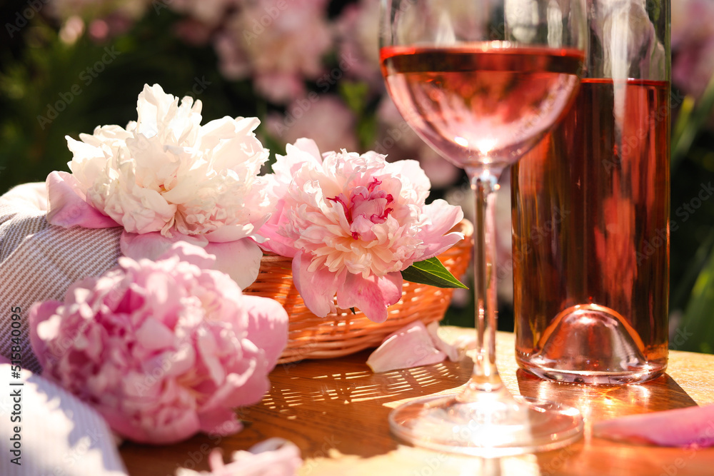 Wall mural Bottle and glass of rose wine near beautiful peonies on wooden table in garden, closeup