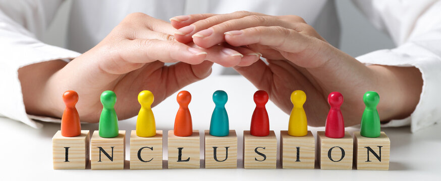 Woman protecting colorful pawns and wooden cubes with word Inclusion at white table, closeup. Banner design
