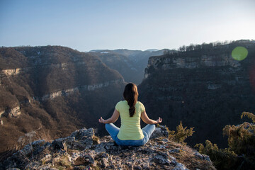 Naklejka na ściany i meble A girl sits on the edge of a cliff in a lotus position, meditates in blue jeans and a yellow T-shirt, view from the back