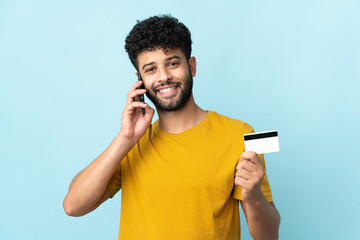 Young Moroccan man isolated on blue background keeping a conversation with the mobile phone and...