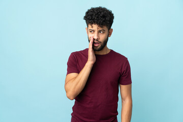 Young Moroccan man isolated on blue background whispering something with surprise gesture while...