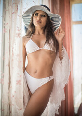 Attractive sexy brunette in white lingerie and big hat posing provocatively in front of a window . Portrait of sensual woman with long hair, ,white bikini and bra in classic boudoir  indoor scene