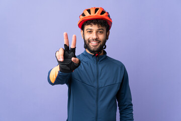 Young Moroccan cyclist man isolated on purple background smiling and showing victory sign - obrazy, fototapety, plakaty