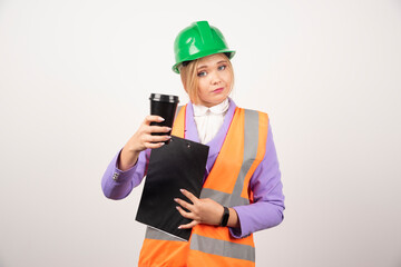 Young female industrial engineer in uniform with clipboard and black cup on white background