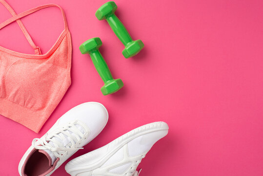 Fitness concept. Top view photo of pink sports bra green dumbbells and white sneakers on isolated pink background