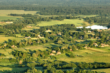 Fototapeta na wymiar Village at lake, aerial view. Country houses at river in countryside. Roofs of country wooden houses near a pond in village. Green farm fields and forests in rural landscape. Farmland, Agriculture.