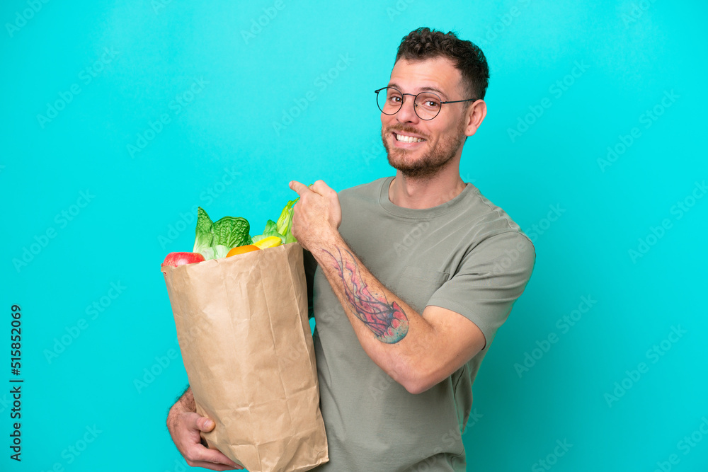 Wall mural Young Brazilian man holding a grocery shopping bag isolated on blue background pointing back