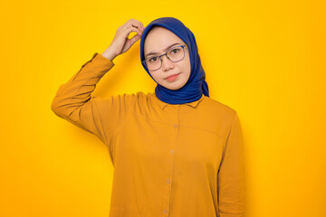 Confused young Asian Muslim woman dressed in orange shirt touching his head and looking with suspicion isolated on yellow background