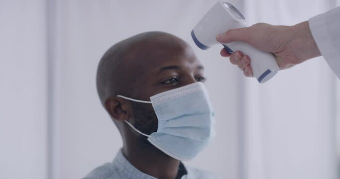 Man Having His Temperature Checked While Wearing A Mask In A Medical Facility By A Health Care Worker During A Pandemic. Young Male Being Quarantined Alone For Isolation During Virus Outbreak.