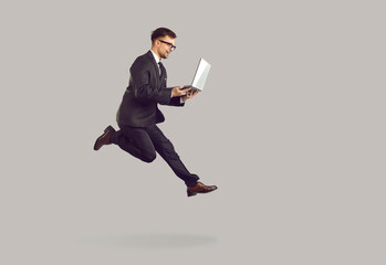 Fototapeta na wymiar Successful joyful young businessman running with laptop in hurry to boost his career. Excited man in suit and glasses runs and jumps with laptop on gray background. Full length. Banner.