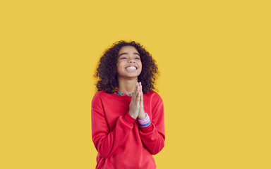 Smiling teen African American girl isolated on yellow studio background keep hands in prayer. Overjoyed funny biracial teenager kid with please or ask hand gesture. Gratitude and faith.