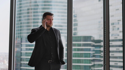 A young man in a business suit is talking on the phone and smiling against the backdrop of a skyscraper. Business concept. Businessman talking on the phone against the backdrop of skyscrapers. 