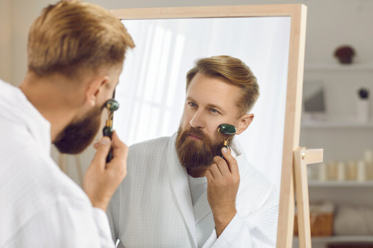 Young Caucasian man use roller massage face for glowing healthy skin. Millennial guy metrosexual do morning beauty procedures in bathroom, have facial treatment. Skincare, cosmetology.