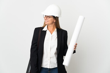 Middle age architect woman with helmet and holding blueprints over isolated background looking to the side and smiling