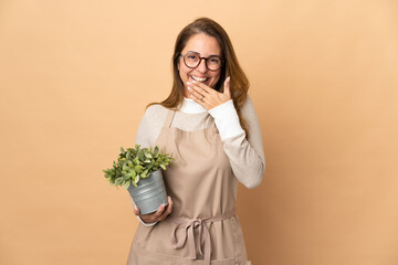 Middle age gardener woman holding a plant isolated on beige background happy and smiling covering mouth with hand