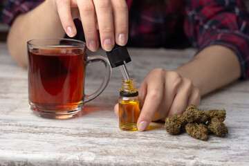 Women's hands are picking up a couple of drops of cannabis cbd oil from a glass bottle into a pipette to add them to tea.  Several dry buds of medical marijuana lie nearby