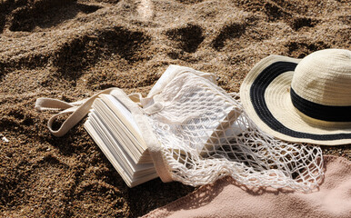 Beach accessories: eco-friendly mesh shopping bag, straw hat, book on sand beach