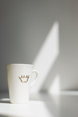 A white tall porcelain coffee cup with a crown on a white table with sun hitting the wall behind creating a nice morning atmosphere Nr.10