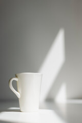 A white tall porcelain coffee cup on a white table with sun hitting the wall behind creating a nice...