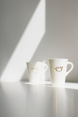 Two white tall porcelain coffee cups with crowns on a white table with sun hitting the wall behind creating a nice morning atmosphere