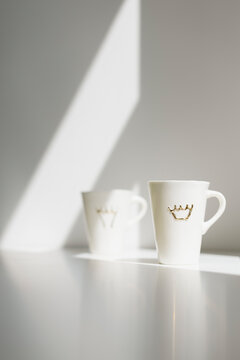 Two White Tall Porcelain Coffee Cups With Crowns On A White Table With Sun Hitting The Wall Behind Creating A Nice Morning Atmosphere